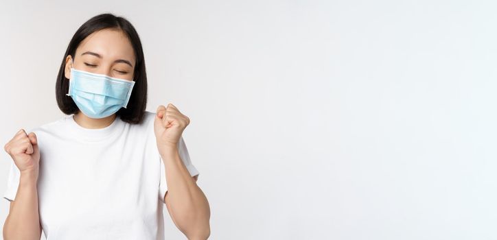 Covid-19, healthcare and medical concept. Enthusiastic asian woman in medical face mask, dancing and celebrating, winning, achieve goal, standing over white background.