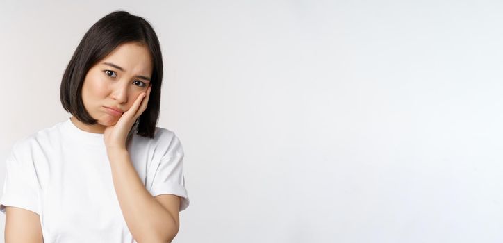 Sad asian girl looking upset and lonely, sulking and frowning, standing against white background in casual tshirt.