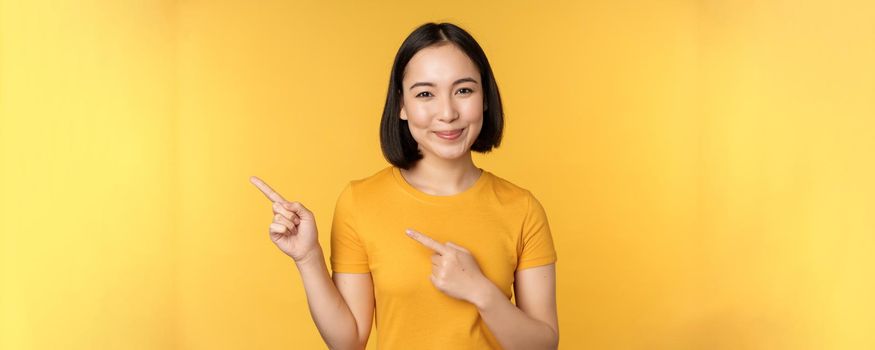 Smiling asian woman pointing fingers left, showing advertisement on empty copy space, standing over yellow background.