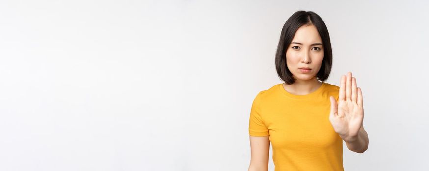 Portrait of asian woman looking serious and angry, showing stop prohibit gesture, taboo sign, forbidding smth, standing in yellow tshirt over white background.