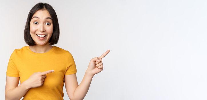 Amazed young asian woman, showing advertisement aside, pointing fingers right at promotion text, brand logo, standing happy against white background.