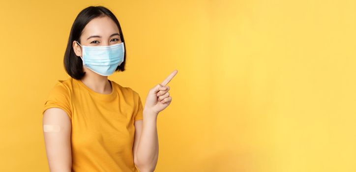 Vaccination from covid and health concept. Image of smiling korean girl in medical face mask, band aid on shoulder, pointing finger at banner advertisement, yellow background.