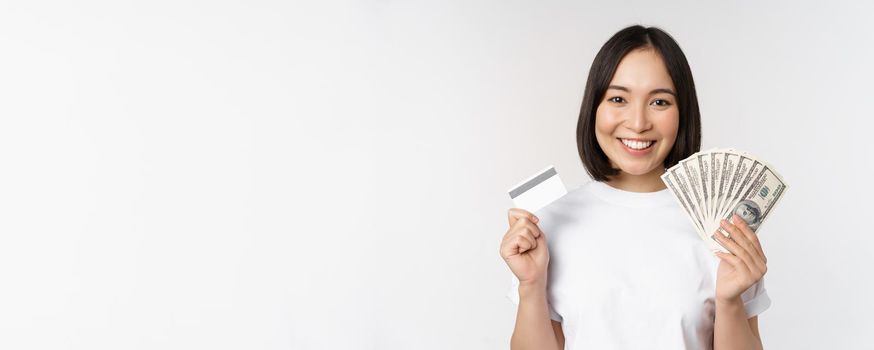 Portrait of asian woman smiling, holding credit card and money cash, dollars, standing in tshirt over white background.