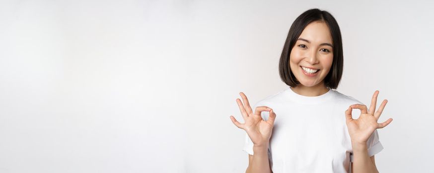 Very well, excellent. Smiling asian woman showing okay sign, approval, ok gesture, looking satisfied, recommending smth, standing over white background.