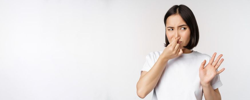 Asian girl looks disgusted, rejecting product with bad smell, shut nose from aversion and cringe, standing against white background.