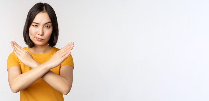 Portrait of asian woman looking serious and angry, showing stop prohibit gesture, taboo sign, forbidding smth, standing in yellow tshirt over white background.