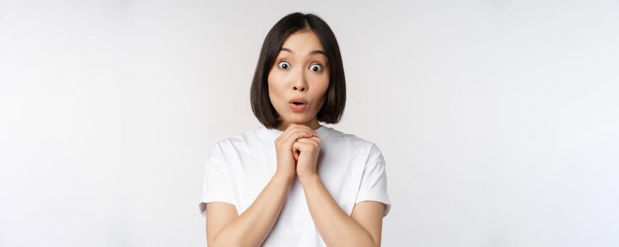 Close up portrait of asian brunette woman looking amazed, say wow, watching smth impressive, standing over white background.