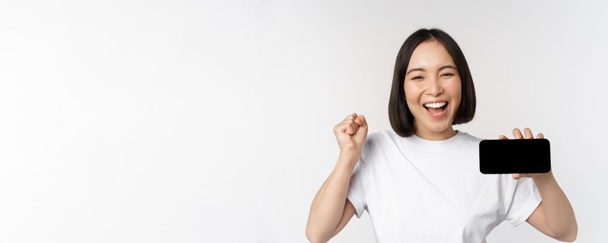 Enthusiastic asian girl scream in joy, showing horizontal smartphone screen, mobile phone display, standing over white background. Copy space
