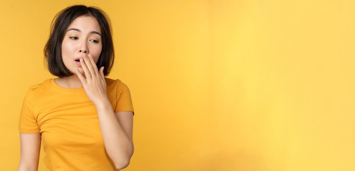 Image of young asian girl student, yawning, feeling tired, standing sleepy agaisnt yellow background. Copy space