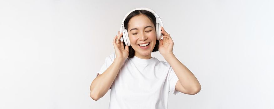 Modern asian girl dancing, listening music with headphones, smiling happy, standing in tshirt over white background.