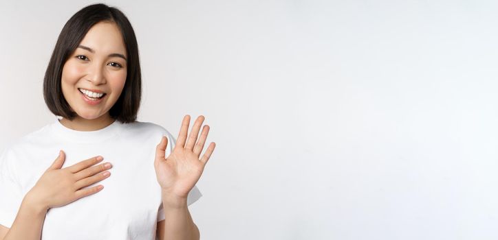 Portrait of beautiful korean girl raising hand, introduce herself, put hand on heart, greeting, standing over white background.