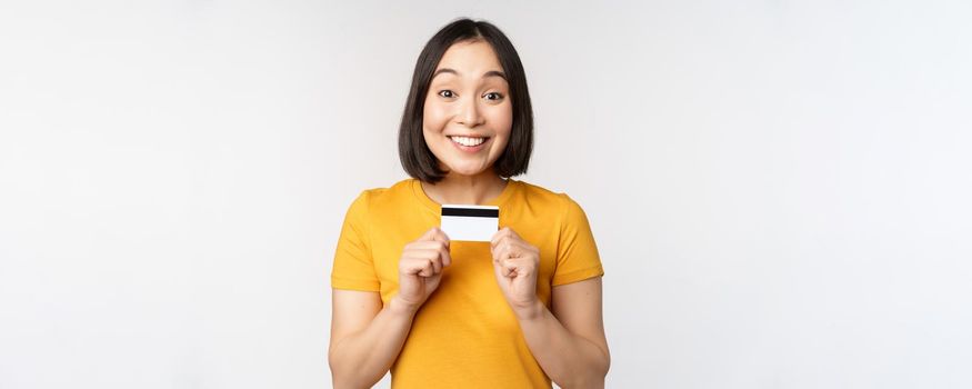 Portrait of beautiful korean girl holding credit card, recommending bank service, standing in yellow tshirt over white background.