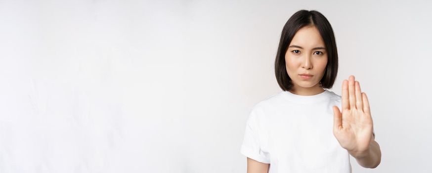 Image of asian girl showing stop, prohibit smth, extend one arm to show forbidding, taboo gesture, standing in tshirt over white background.