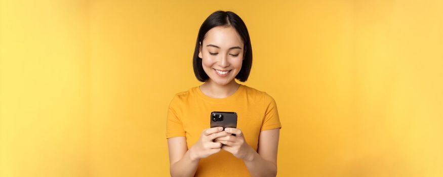 Technology. Smiling asian woman using mobile phone, holding smartphone in hands, standing in t-shirt against yellow background.