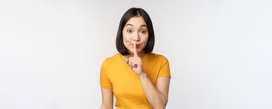 Keep quiet. Cute asian woman make shhh gest, showing shush, hush sign, press finger to lips, silence, standing over white background.