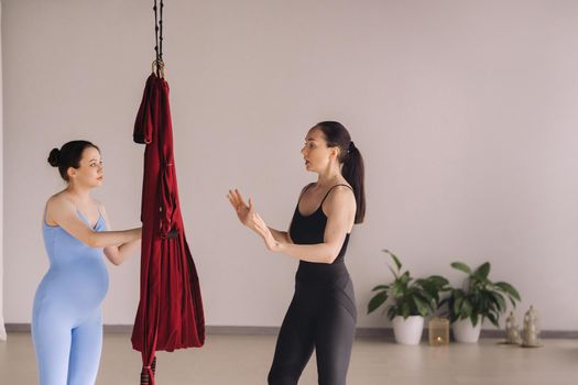 Pregnant girl. A woman with a trainer does yoga in a hammock in the gym. The concept of a healthy lifestyle, motherhood.