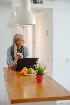 Joyful lady standing by kitchen table and holding cup of hot drink while using modern notebook