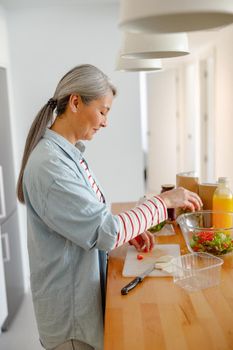 Female person standing by wooden table and putting tomatoes in bowl with salad while cooking at home