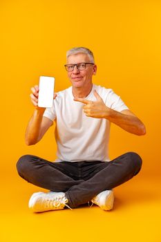 Senior handsome smiling man holding smartphone showing screen over yellow background, close up
