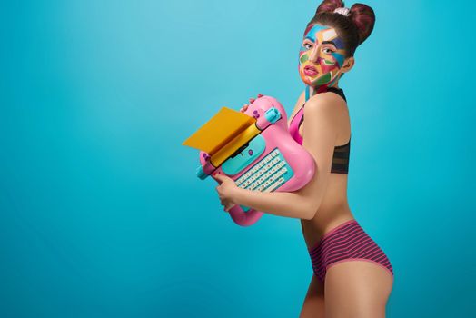 Skittish girl holding pink toy typewriter, looking at camera with seductive look, opened mouth. Attractive, cool girl posing, has funky pop art make up and bow hairstyle.