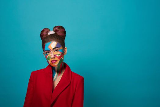 Stylish, trendy model wearing in red jacket, posing in studio with blue background. Cool girl has creative pop art make up on face and nice bow hairstyle. Funky lady looking at camera, posing.