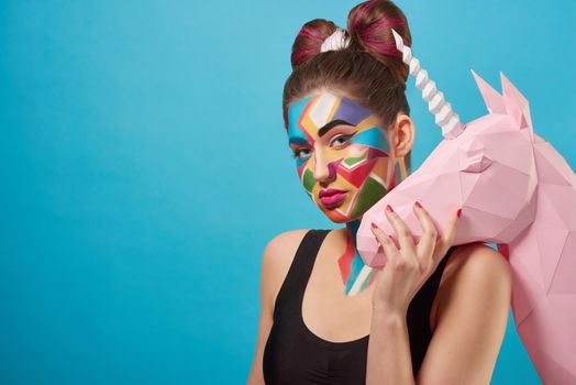 Portrait of beautiful model posing, looking at camera. Charming woman holding hand on 3D paper unicorn head. Funky girl has pop art creative make up. Papercraft concept.
