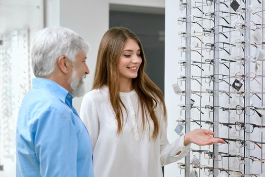 Sideview of smiling assistant showing eyeglasses to old patient. Advising which optical glasses are better. Tranparent stand with glasses set in front of white wall on background. friendly and kind.