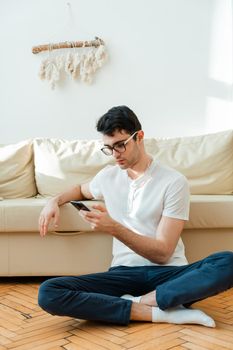 Portrait of an attractive, smiling man in casual clothes sitting on the floor near the sofa in the living room and watching the news using a mobile phone