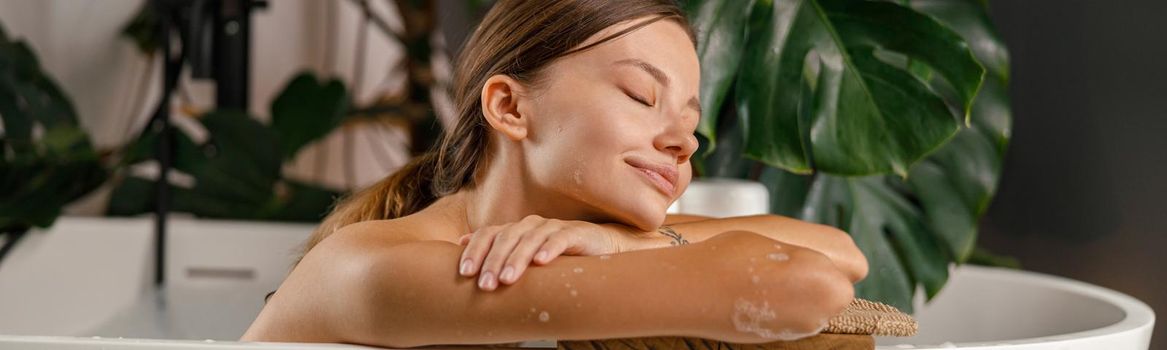 Dreamy young woman leaning on bathtub side and resting in the bathroom decorated with tropical plant. Wellness, beauty and care concept