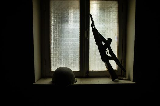 Conceptual photo of war between Russia and Ukraine. Russian weapon and helmet on windowsill at night. Old creepy room with window. Explosion outside.