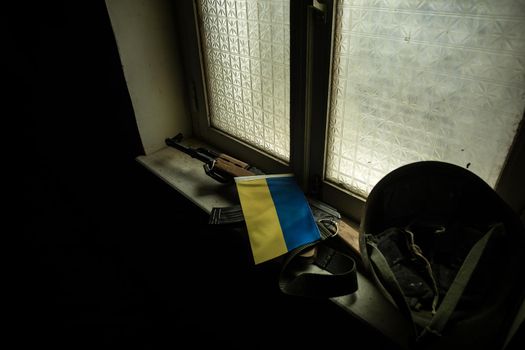 Conceptual photo of war between Russia and Ukraine. Ukrainian flag, helmet and gun on windowsill at night. Old creepy room with window. Explosion outside.