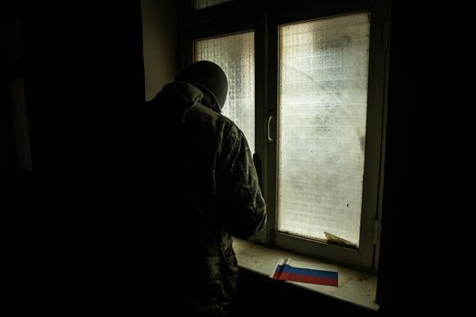 Conceptual photo of war between Russia and Ukraine. Silhouette of a russian soldier with gun at window with russian flag on windowsill. Old creepy room with window. Explosion outside.