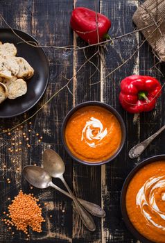 Delicious pumpkin soup with heavy cream on dark rustic wooden table with red bell pepper, bread toasts, lentil and autumn branches. Top view