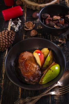 Roasted duck breast with golden crispy skin served with baked apples and chestnuts. Served on a ceramic black plate over rustic dark wooden table with a deer napkin ring and candles. Close up