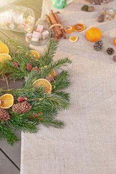 Rustic Christmas table with natural spruce wreath, dried oranges, fresh tangerines, pine cones, cinnamon sprigs and sweets on a fabric tablecloth. Copy space. Vertical