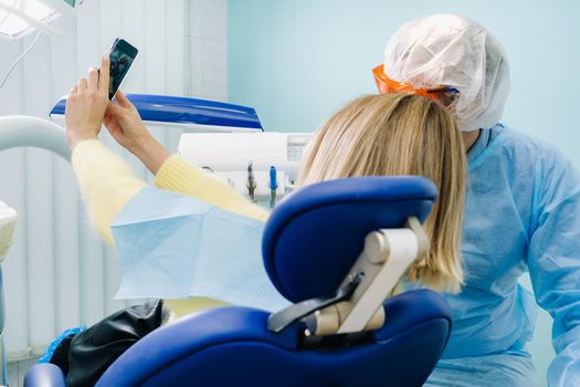 a dentist in a protective mask sits next to a patient and takes a selfie photo while working.