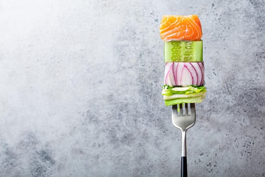 Close-up of fork with food on it: delicious fillet salmon, cucumber, onion, green salad on gray stone rustic background. Concept of healthy diet and clean eating, balanced nutrition space for text