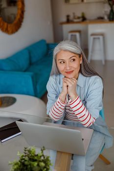 Cheerful female person looking away and smiling while working on notebook in living room