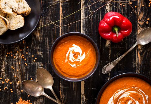 Delicious pumpkin soup with heavy cream on dark rustic wooden table with red bell pepper, bread toasts, lentil and autumn branches. Top view