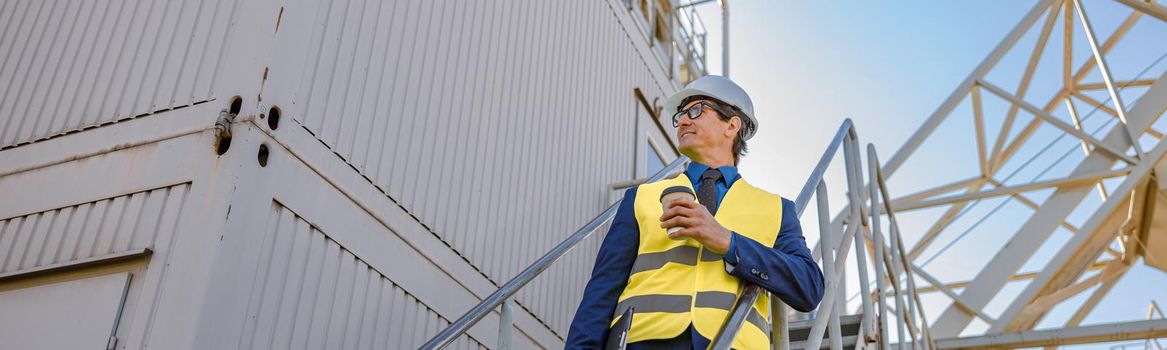 Smiling man factory worker holding takeaway drink and folder while standing on staircase outside industrial building