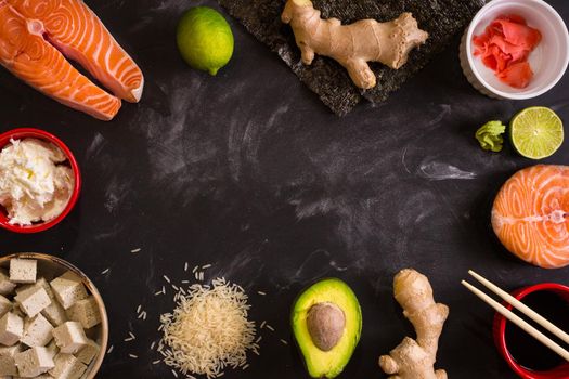 Overhead shot of ingredients for sushi on dark background. Raw salmon steak, rice, cream cheese, avocado, lime, pickled ginger (gari), raw ginger, wasabi, soy sauce, nori, сhopsticks. Asian food background. Space for text....
