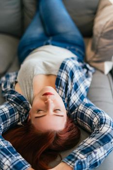 Young red-haired girl in grey T-shirt, blue and white checked shirt and jeans relaxing on the sofa. Vertical body shot while resting from a day's work.