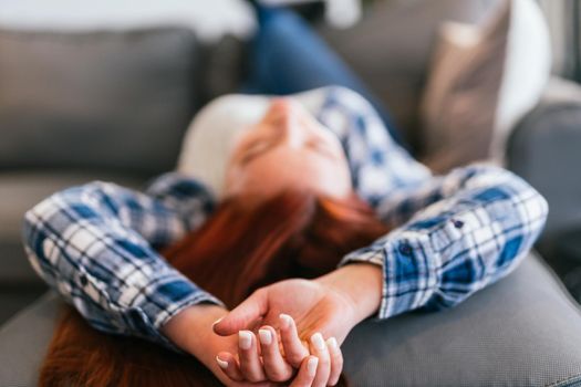 Detailed plan of the hands of young red-haired girl in grey T-shirt, blue and white checked shirt and jeans relaxing on the sofa. Vertical body shot while resting from a day's work.