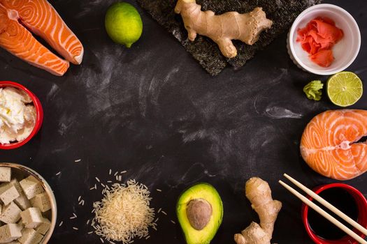 Overhead shot of ingredients for sushi on dark background. Raw salmon steak, rice, cream cheese, avocado, lime, pickled ginger (gari), raw ginger, wasabi, soy sauce, nori, сhopsticks. Asian food background. Space for text....