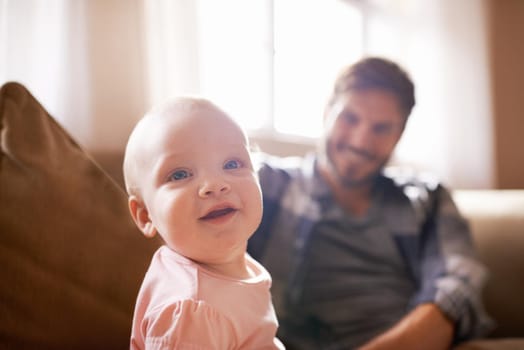 Shot of an adorable baby girl sitting on the sofa with her father in the background.