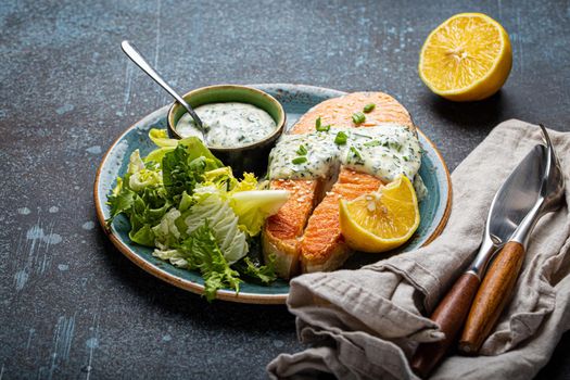 Healthy food meal cooked grilled salmon steak with white dill sauce and green salad leafs on plate on rustic concrete stone background table angle view, diet healthy nutrition dinner