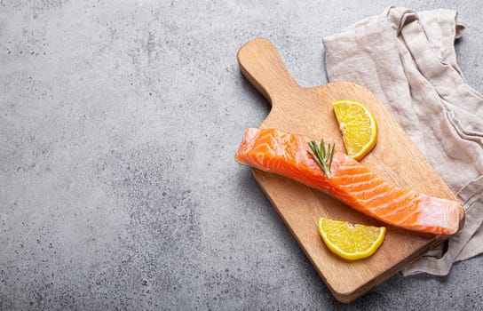 Raw salmon fish fillet with lemon wedges and rosemary on wooden cutting board on gray stone concrete rustic background kitchen table from above, healthy eating and diet, space for text