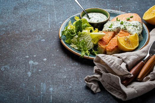 Healthy food meal cooked grilled salmon steak with white dill sauce and green salad leafs on plate on rustic concrete stone background table angle view, diet healthy nutrition dinner copy space