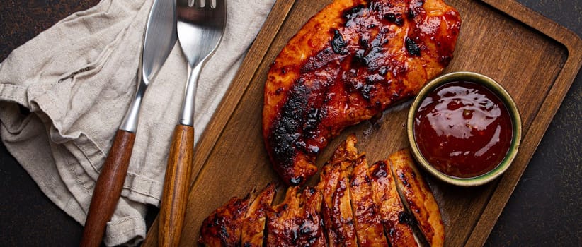 Grilled turkey or chicken marinated fillet with red sauce served and sliced on wooden cutting board on stone brown background from above, poultry breast barbecue