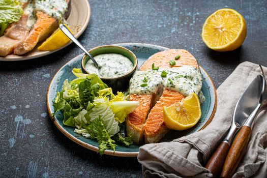 Healthy food meal cooked grilled salmon steak with white dill sauce and green salad leafs on plate on rustic concrete stone background table angle view, diet healthy nutrition dinner